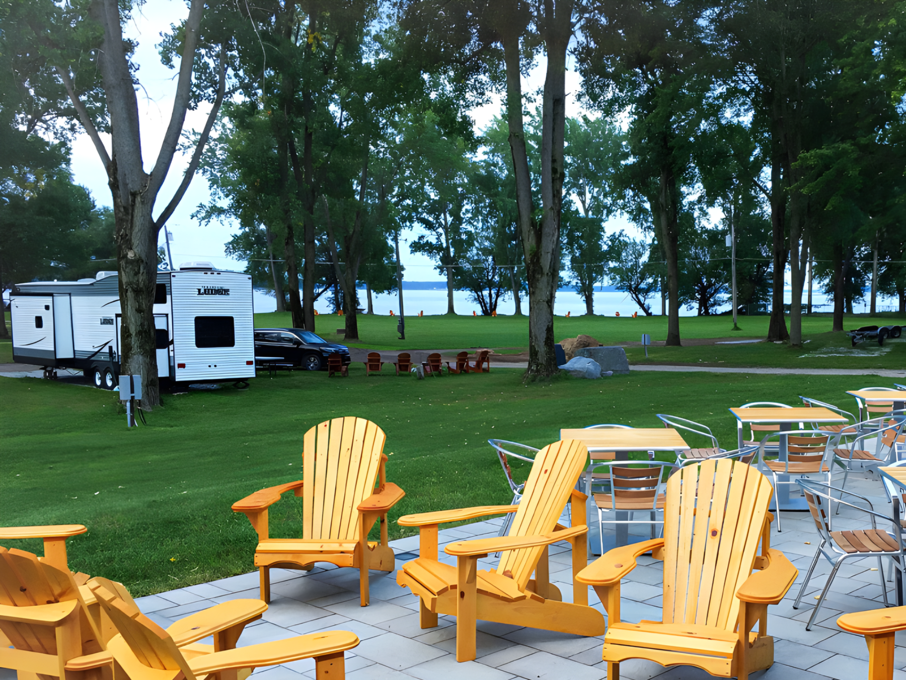 Lakeside scenery at Lake Champlain, NY featuring bright Adirondack chairs, shaded green lawn, RV campsite, and calm water visible through trees.