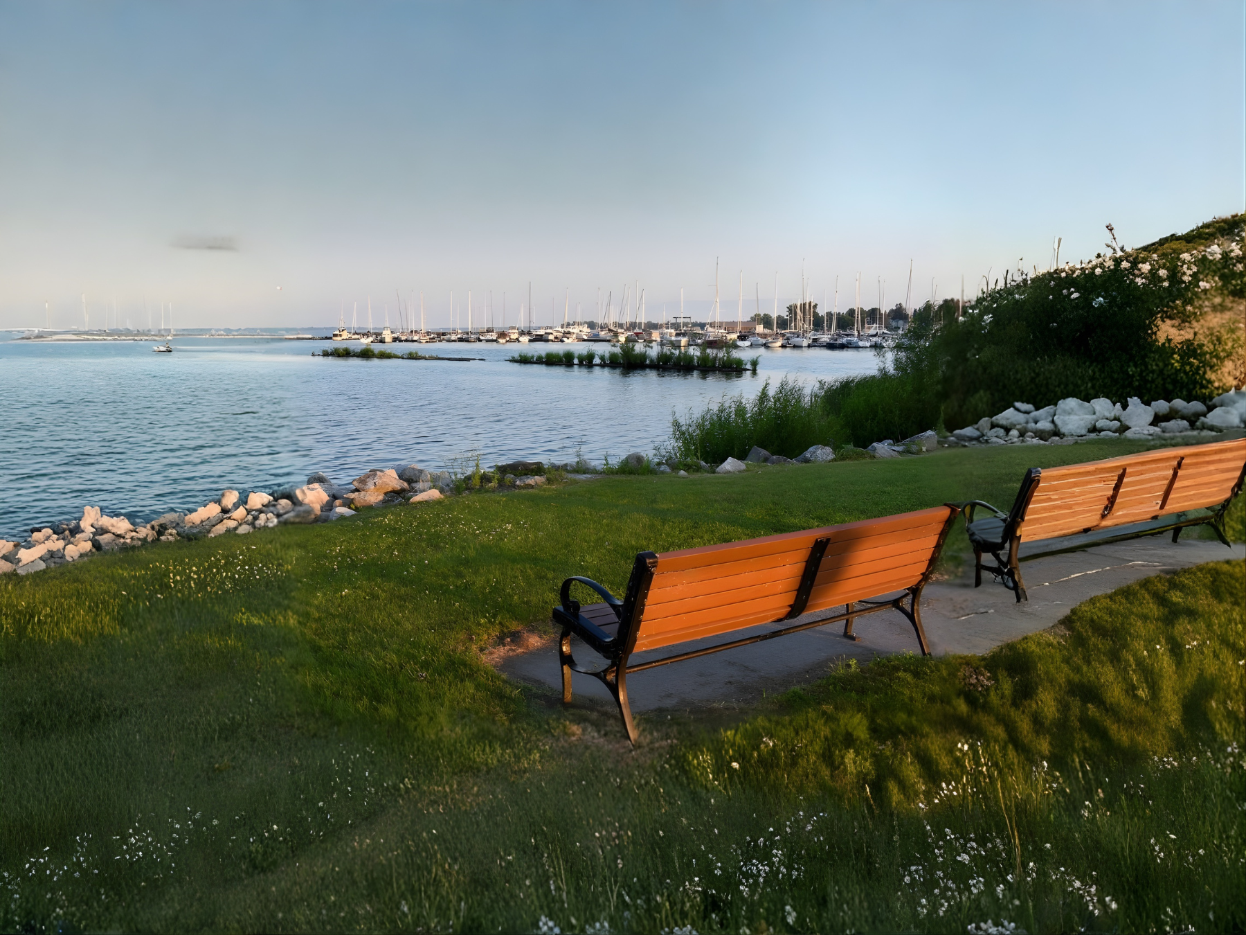 Lakeside scenery at Lake Champlain, NY with benches overlooking calm water and a marina of sailboats.