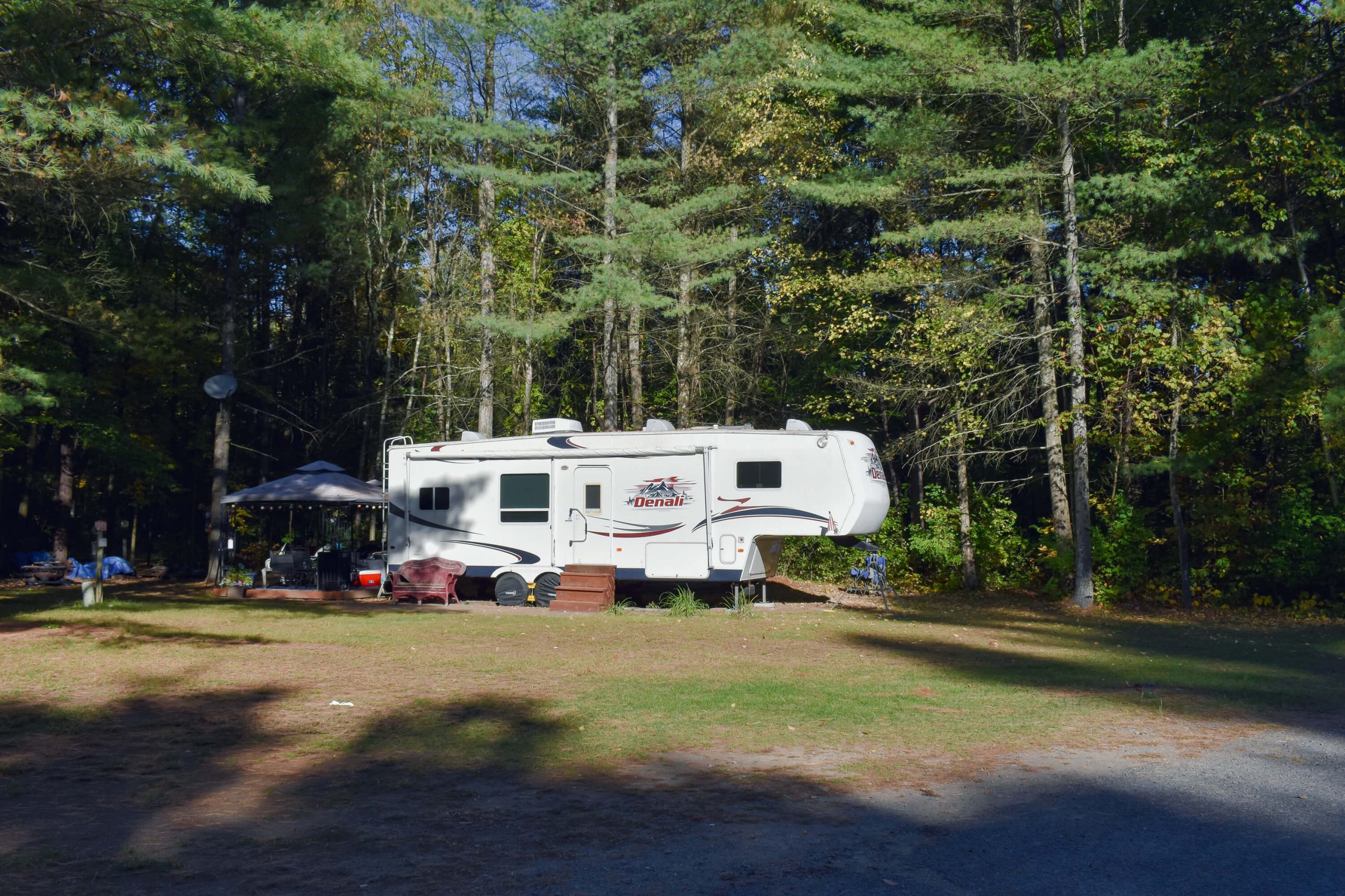 Fifth wheel trailer with canopy setup on a spacious wooded campsite surrounded by tall pine and deciduous trees at a campground in Peru, NY