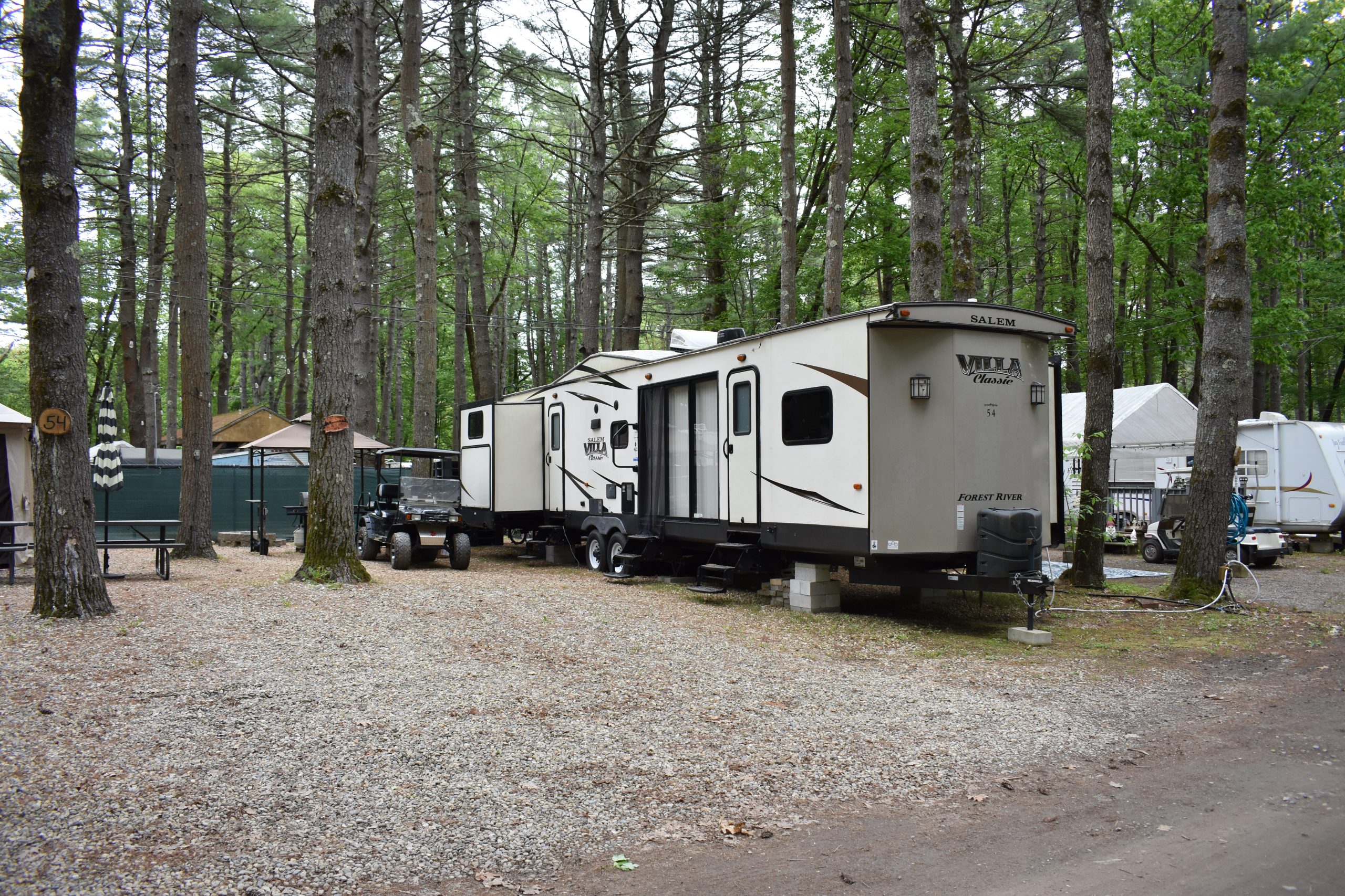 Travel trailers parked at wooded campsites with tall pine trees and shaded gravel pads at a campground near Boston in Templeton, MA