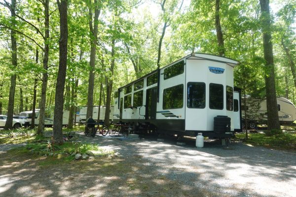 Large destination travel trailer with picture windows parked on a shaded wooded site surrounded by tall trees at a campground in Bellingham, MA