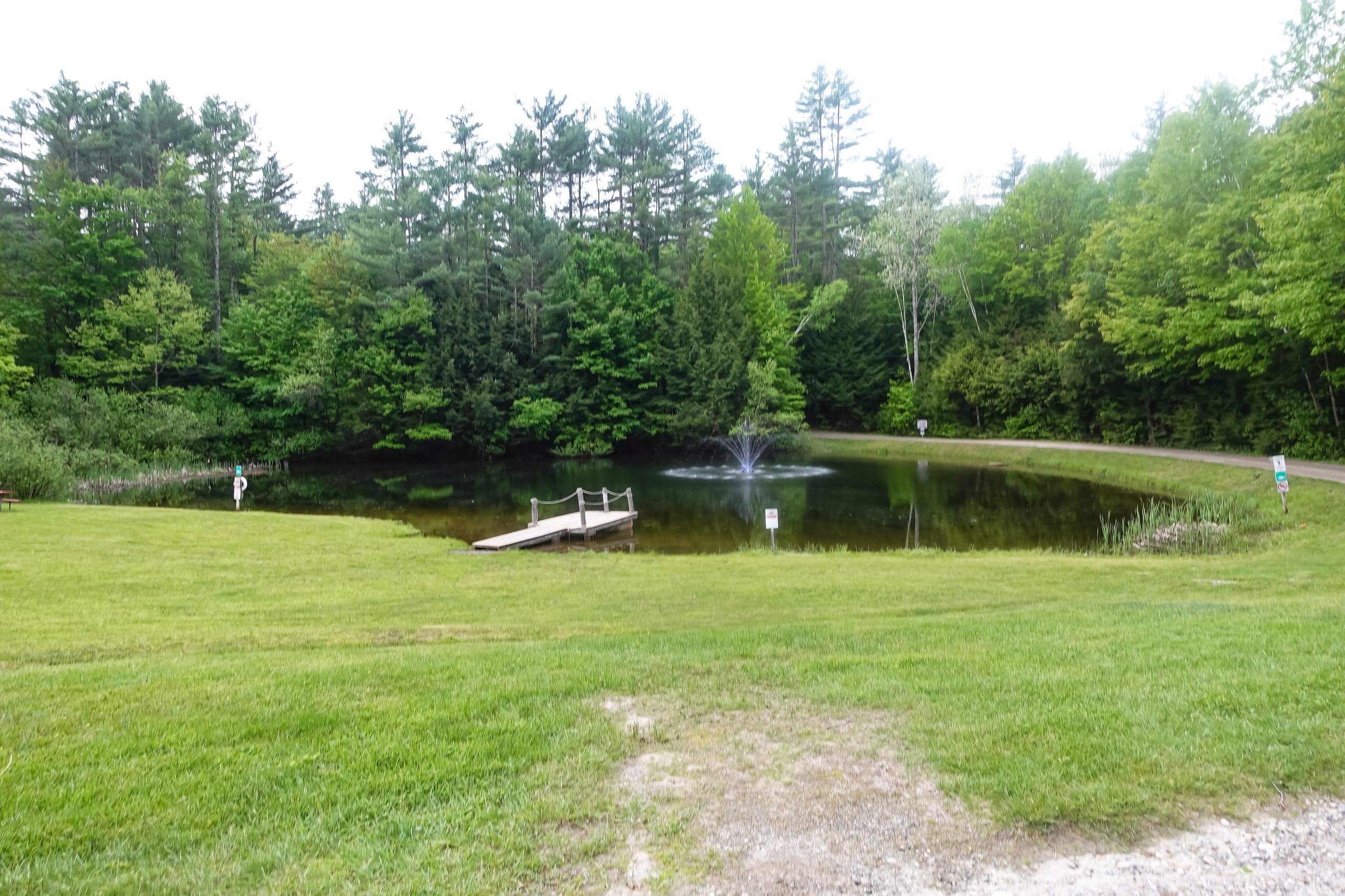 Serene campground pond with a fountain and wooden dock set among lush green grass and evergreen trees at a campground near Mount Sunapee