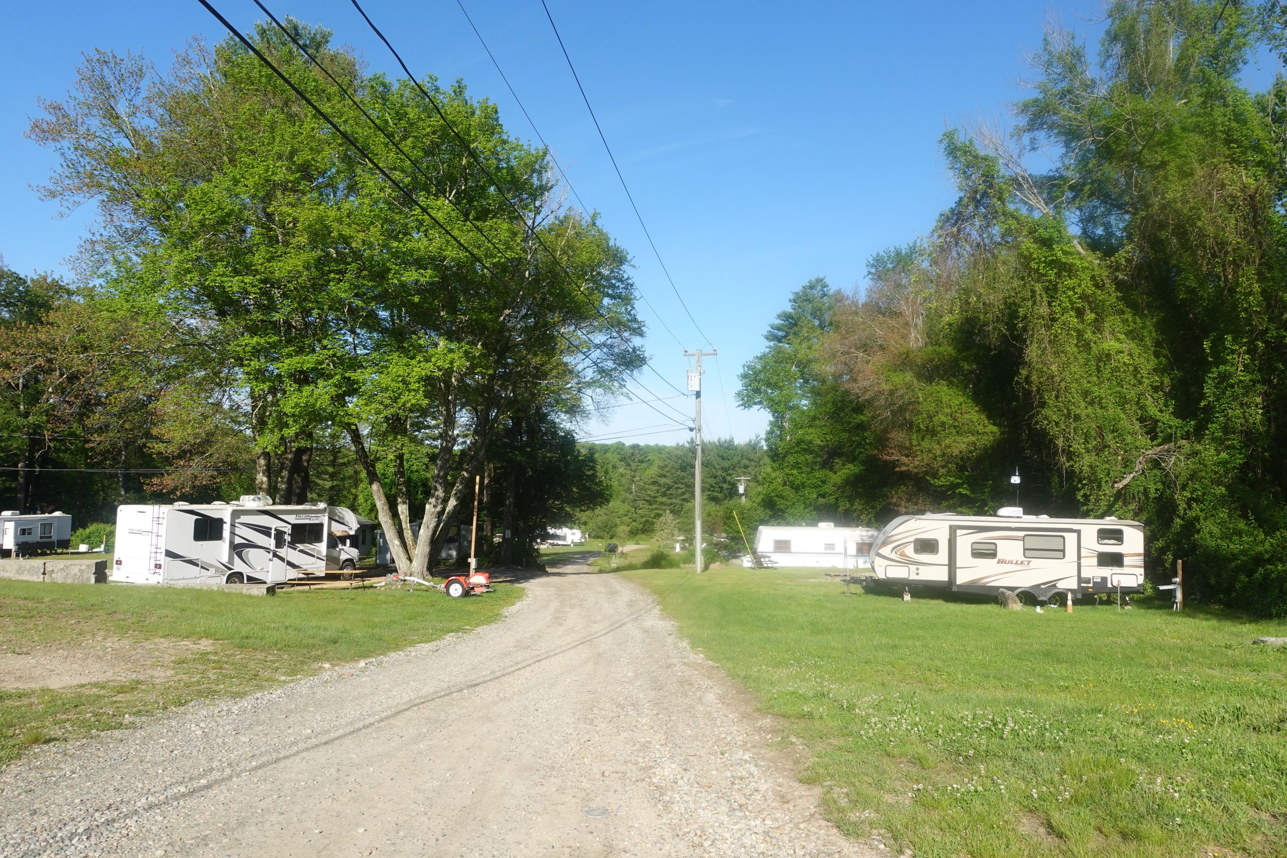 Gravel road leading through open RV and trailer sites with green lawns and mature trees under a clear blue sky at a campsite in Sterling, CT