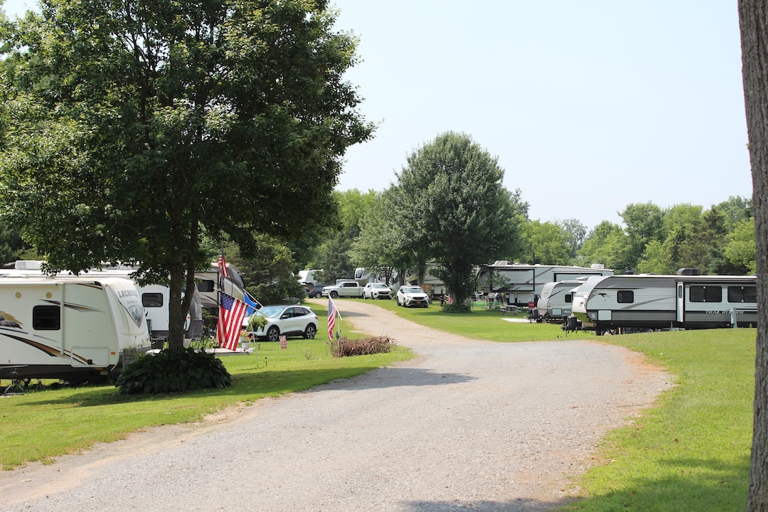 Tree-lined gravel road through RV and travel trailer sites with American flags and green lawns at a campground in Blossvale, NY