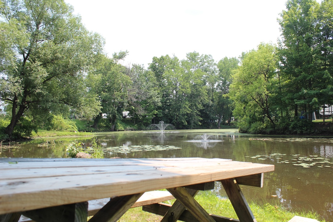 Picnic table overlooking a tranquil pond with a water fountain surrounded by lush green trees at a campground in Blossvale, NY