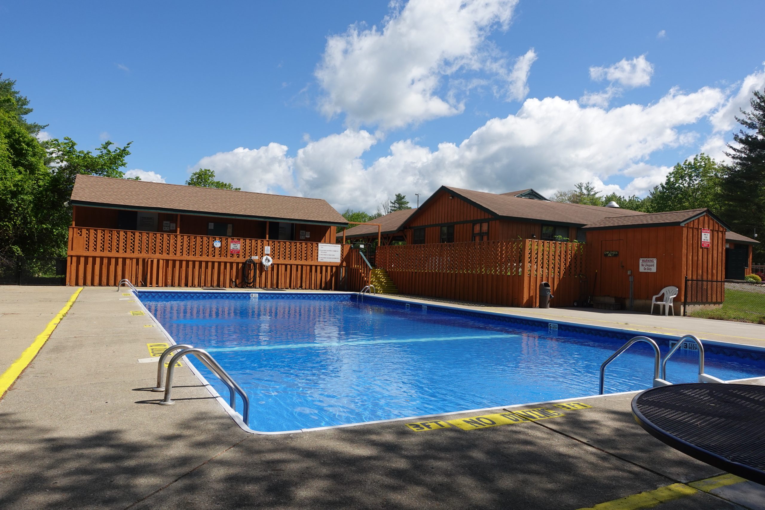 Outdoor swimming pool at White Pines Campsites featuring clear blue water, concrete sun deck, safety ladders, and rustic wooden cabin surroundings on a sunny day.