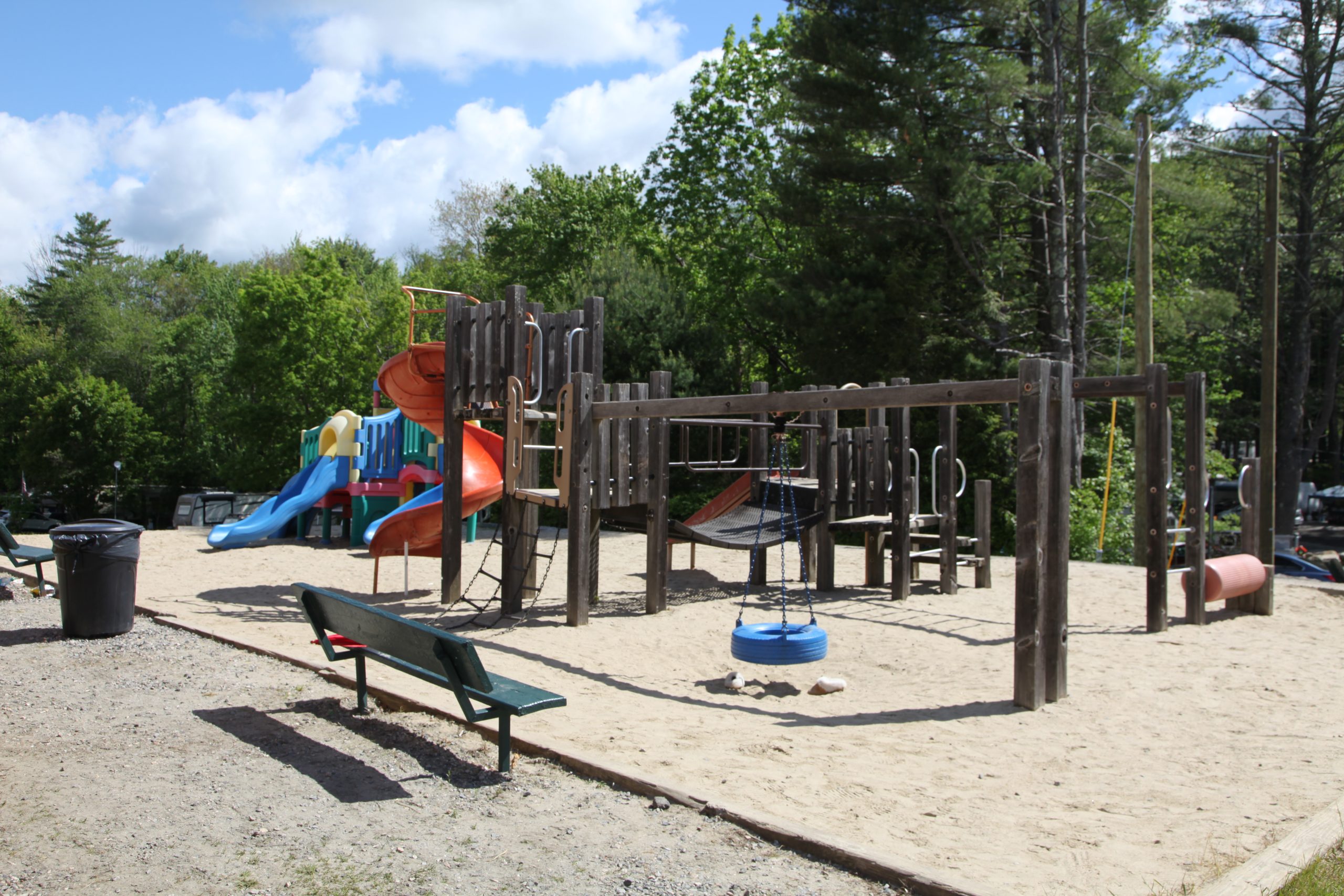 Kids' playground with wooden monkey bars, colorful slides, tire swing, and bench seating on a sandy play area at a campground in Barkhamsted, CT