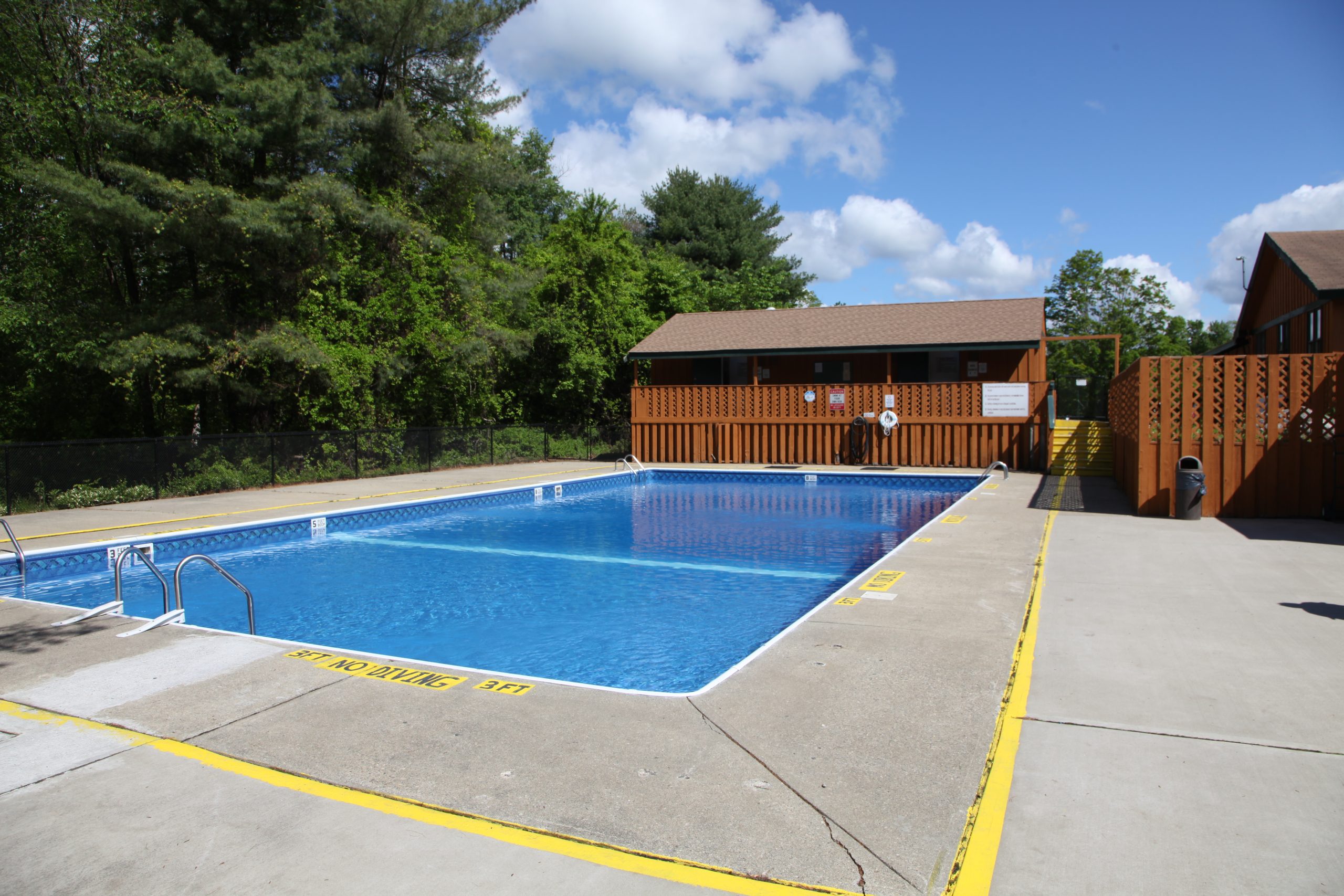 Large outdoor swimming pool with concrete deck and wooden fencing surrounded by trees on a sunny day at a campground in Barkhamsted, CT