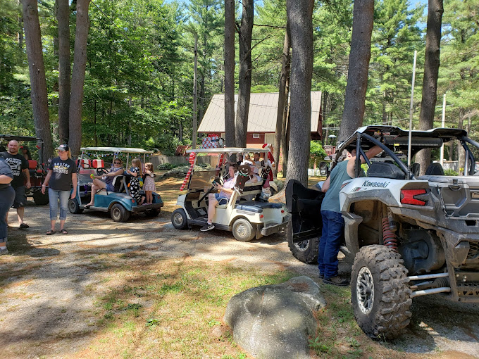 Families enjoying a festive golf cart parade with decorated carts and a Kawasaki UTV under tall pine trees during a community event at a campground near Boston in Templeton, MA