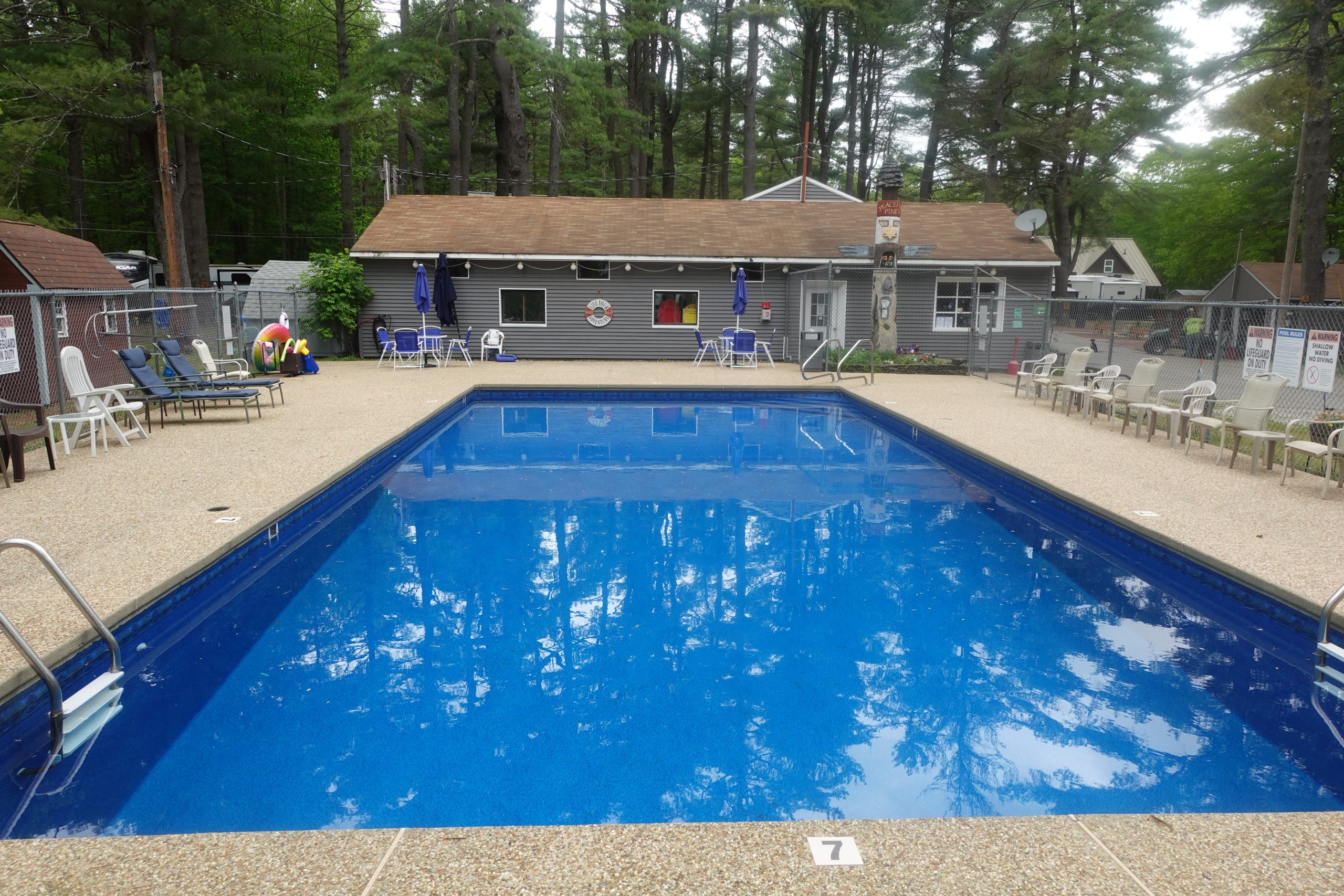 Large in-ground swimming pool with clear blue water surrounded by lounge chairs, pool floats, and a gray clubhouse building nestled among tall pine trees at a campground near Boston in Templeton, MA