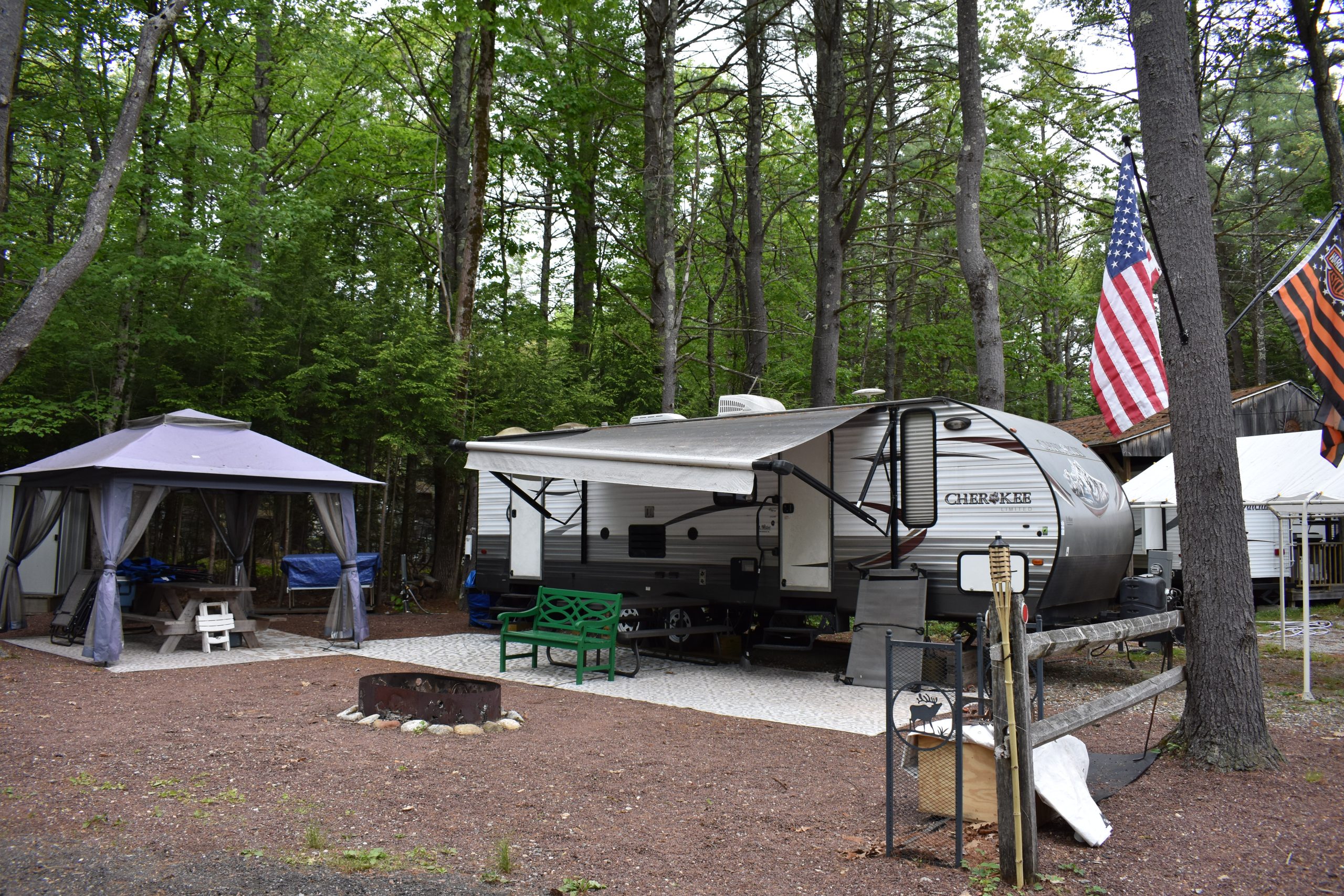 Cherokee travel trailer parked at a wooded campsite in Templeton, MA, featuring a covered gazebo with picnic table, fire pit, green bench, and American flag surrounded by lush green trees at a campground near Boston.