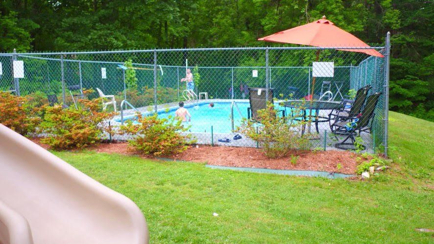 A fenced-in campground swimming pool with lounge chairs and an orange umbrella, surrounded by grass, shrubs, and trees.