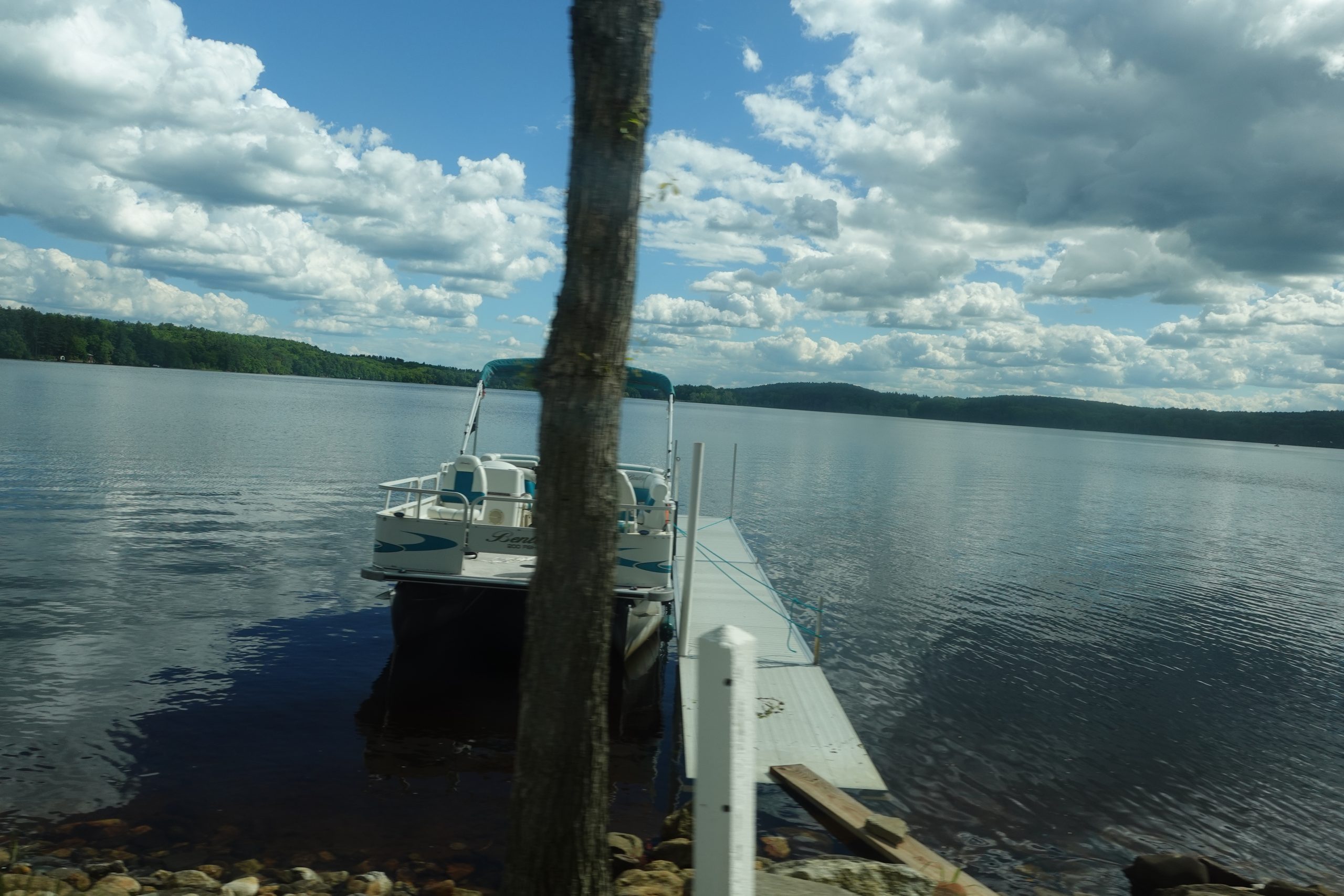 A pontoon boat tied to a small dock on a calm lake with forested shorelines and fluffy clouds overhead.