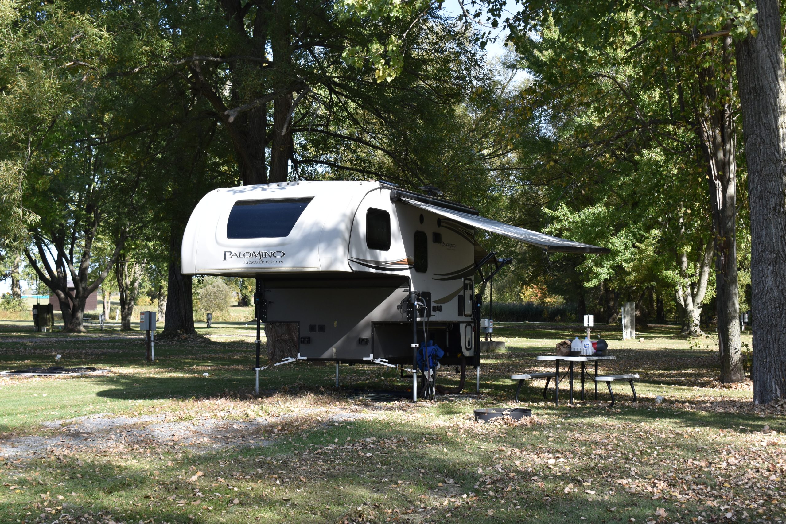 A truck camper set up with an awning at a shaded campsite scattered with fallen autumn leaves.