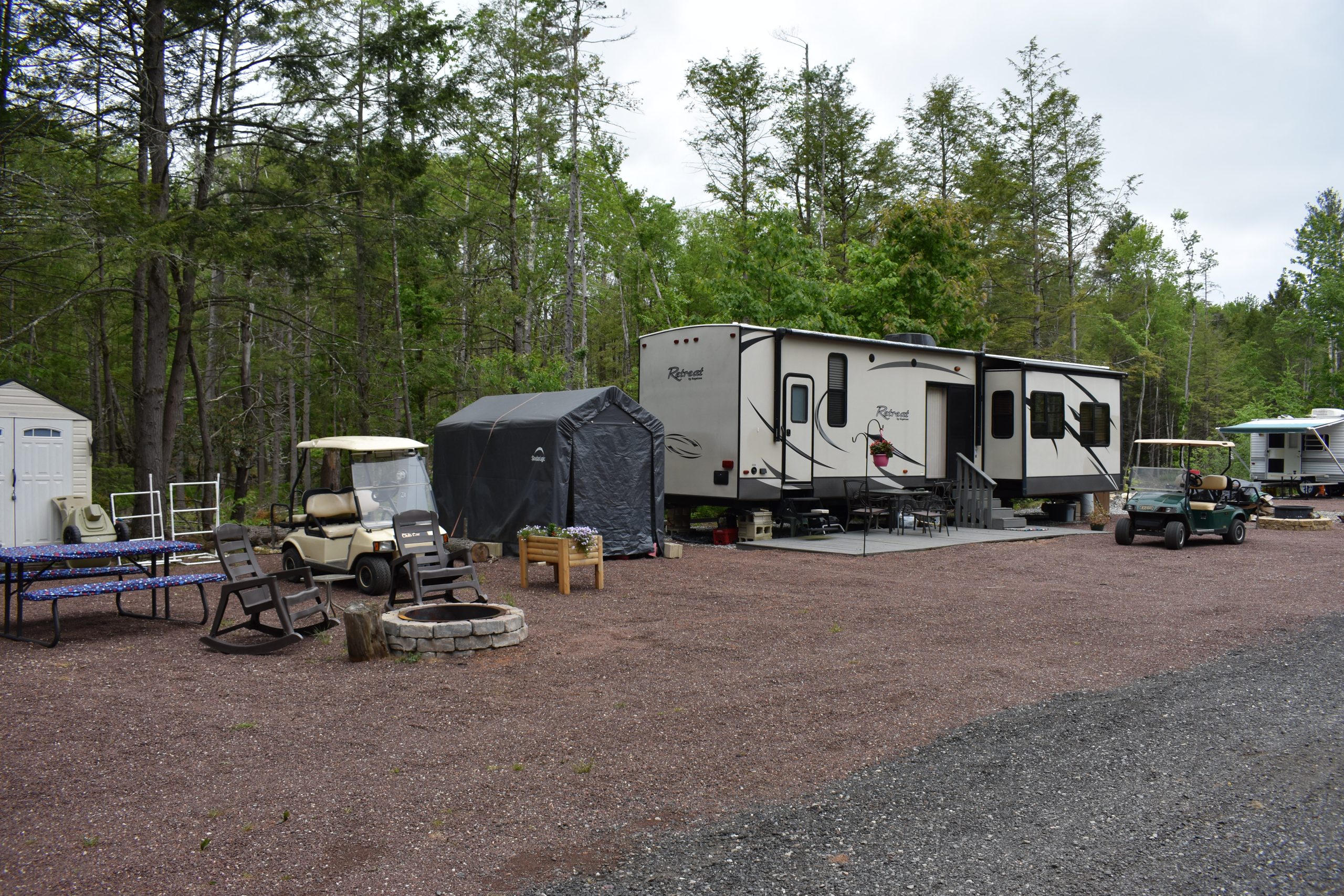 Park Trailer at a seasonal campsite with golf carts, a stone fire pit, rocking chairs, and a storage shelter surrounded by trees.