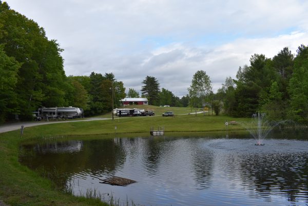 A campground pond with a fountain in the foreground and RVs parked on a grassy hillside near a red barn under cloudy skies.