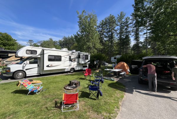 A sunny RV campsite with a Sunseeker motorhome, colorful camp chairs around a fire pit, and a small orange tent under tall trees.