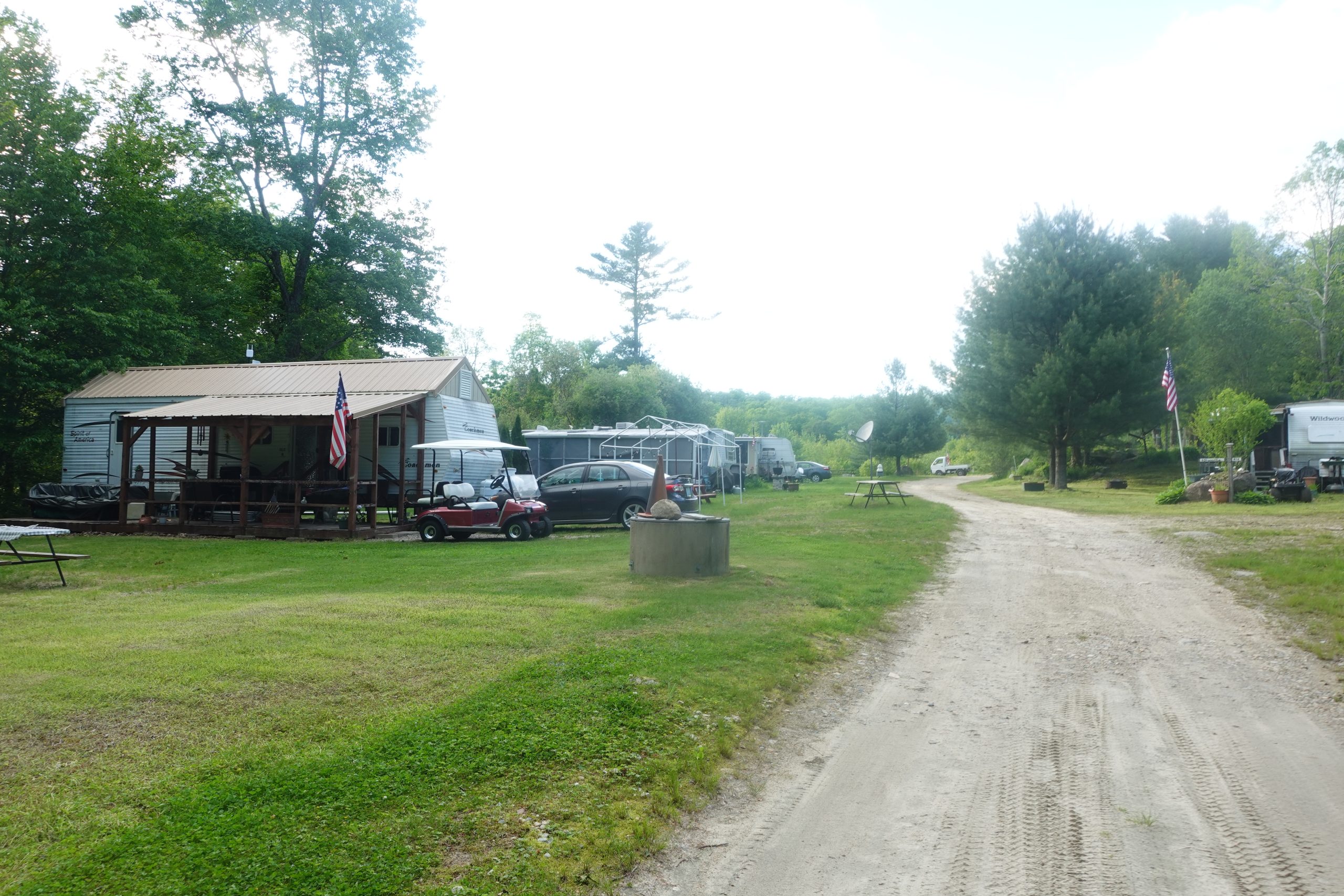 A campground road lined with seasonal RV sites featuring covered porches, a golf cart, and American flags.