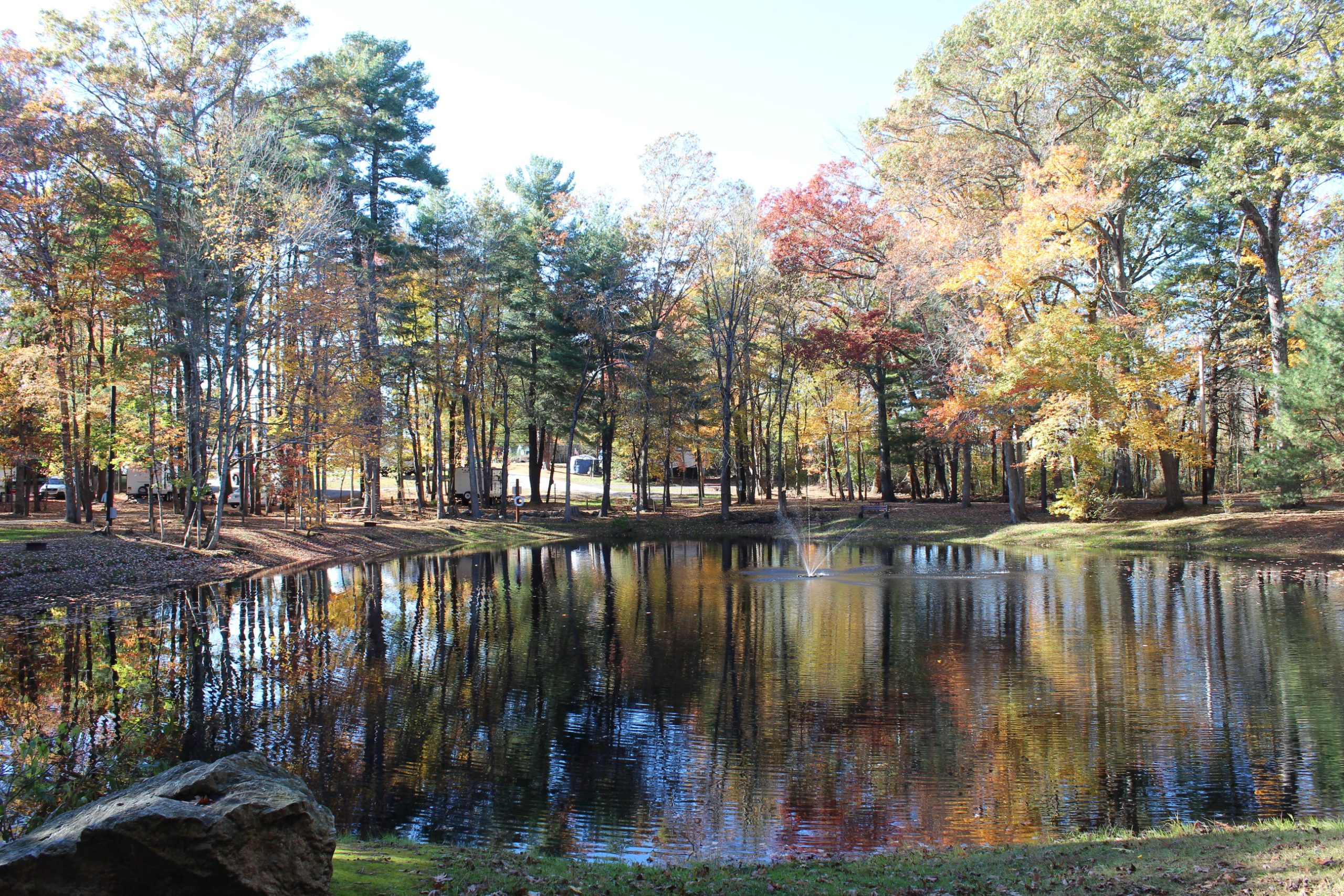 A tranquil pond with a small fountain reflecting colorful autumn trees at a wooded campground.