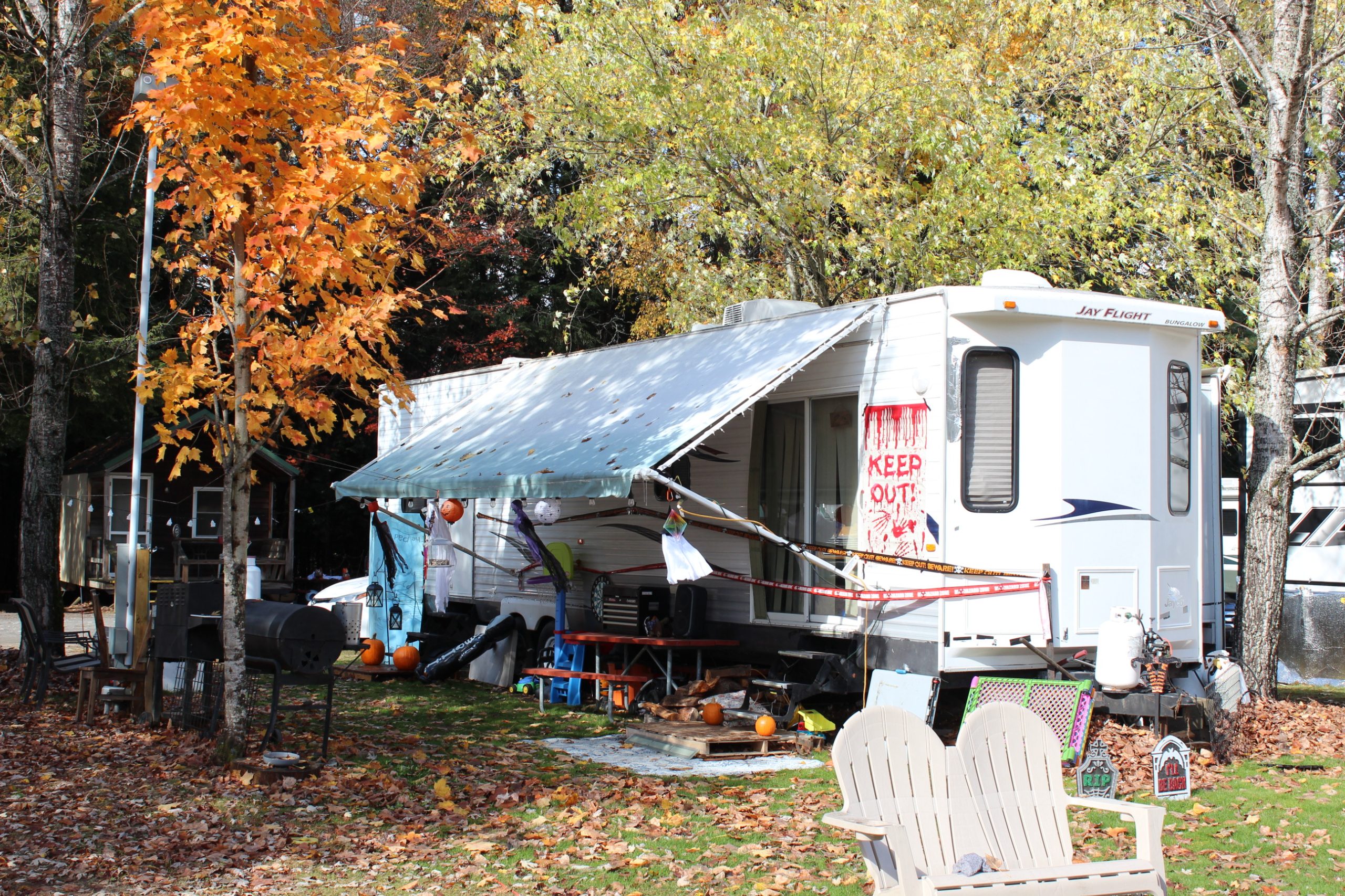 Travel trailer decorated for Halloween with pumpkins, a "Keep Out" sign, and caution tape, set among trees with bright autumn foliage.