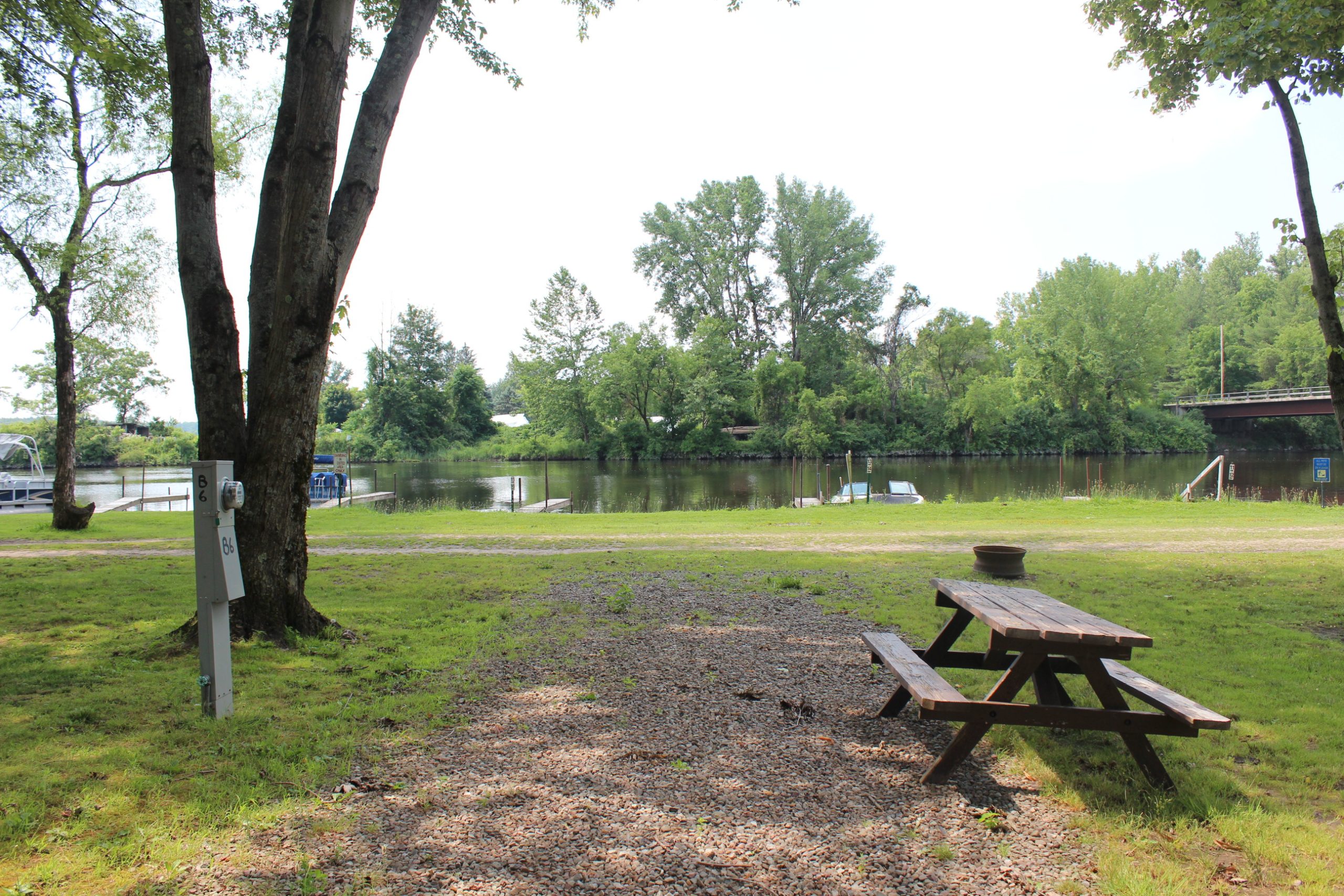 aterfront campsite with a wooden picnic table and fire pit overlooking a calm river lined with trees.