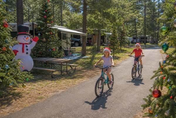 Kids in Santa hats biking past decorated trees and an inflatable snowman during Christmas in July at a campground.