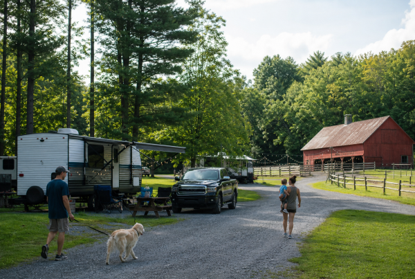 Family walking their dog past RV sites at a farm campground with a red barn and string lights.