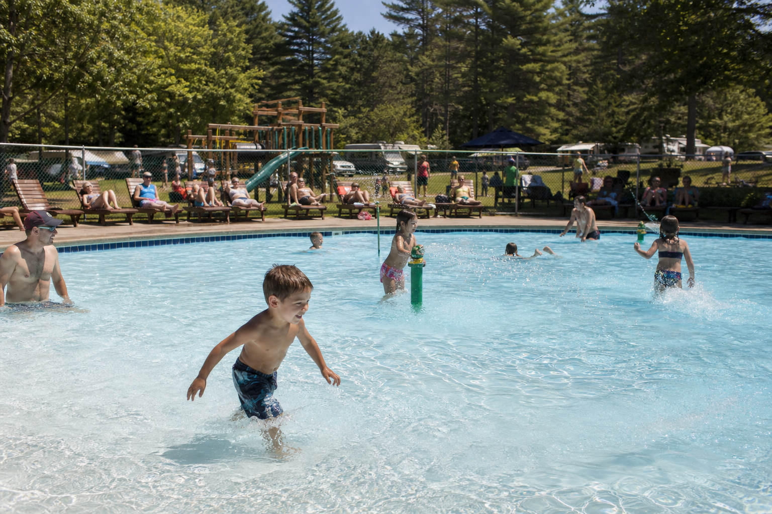 Kids splashing in a campground swimming pool with a playground and RV sites in the background.