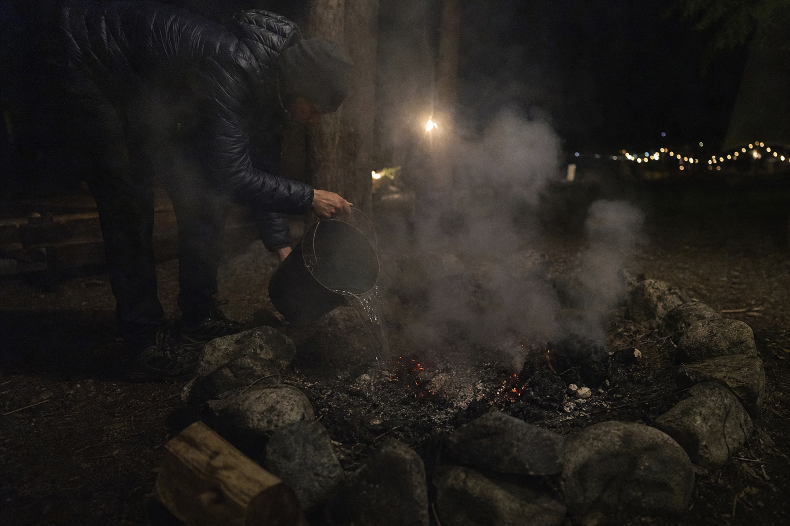 Man pouring a bucket of water over glowing campfire embers at night, sending up a cloud of steam and smoke.