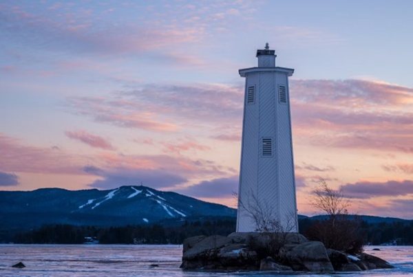 White lighthouse on frozen Lake Sunapee with Mount Sunapee in the background at sunset.